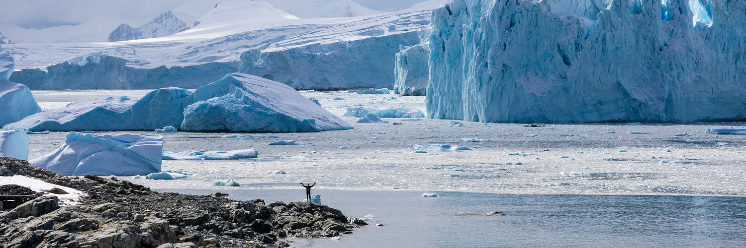 Person standing on rocky shoreline looking toward large Antarctic icebergs and glacier landscape